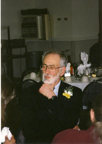 Nat Hart, gray hair, beard, glasses, dark suit jacket with a flower corsage seated at the Faculty and Staff Recognition Dinner in 2001.